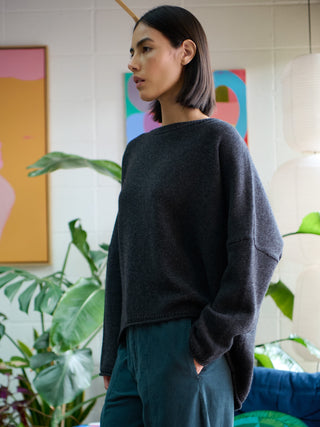 A woman wearing a dark charcoal gray knit sweater and teal pants, standing in a brightly lit room with potted plants and colorful wall art.